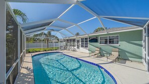 A covered swimming pool area with a clear blue pool, a covered patio with chairs, and a view of a house in the background.