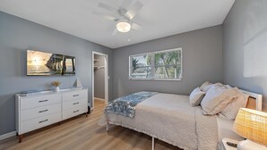 Ein Schlafzimmer mit Bett, Nachttisch, Kommode, Deckenventilator, Fenster mit Jalousien und einem Gemälde an der Wand.