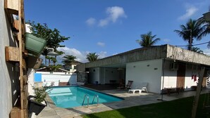 A swimming pool with a stone deck, surrounded by a grassy yard and a building with a flat roof. - Pousada Américas (Guarujá)