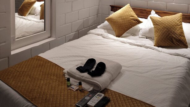 A hotel room with a white brick wall, a bed with white bedding and two brown pillows, a wooden headboard, a mirror, and a bedside table with slippers and toiletries. - Srikrishna Paradise (Tiruvannamalai)