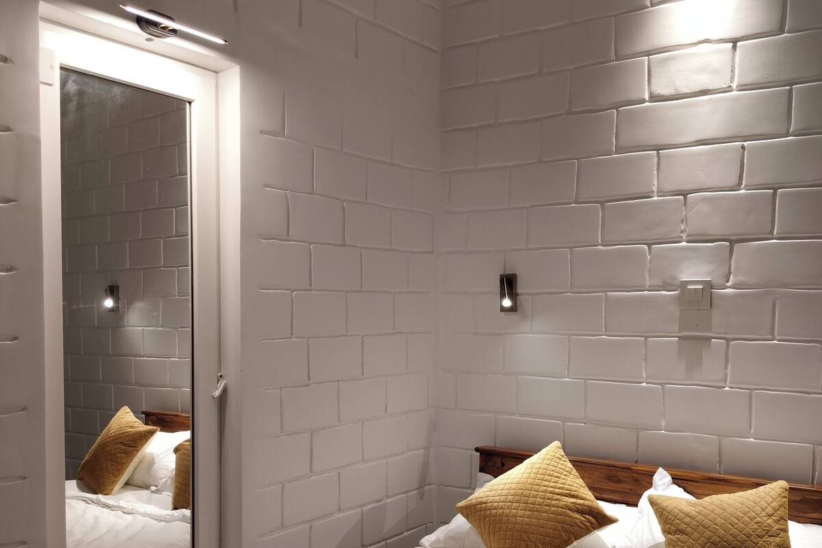 A hotel room with a white brick wall, a bed with white bedding and two brown pillows, a wooden headboard, a mirror, and a bedside table with slippers and toiletries.