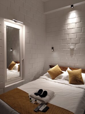 A hotel room with a white brick wall, a bed with white bedding and two brown pillows, a wooden headboard, a mirror, and a bedside table with slippers and toiletries. - Srikrishna Paradise (Tiruvannamalai)