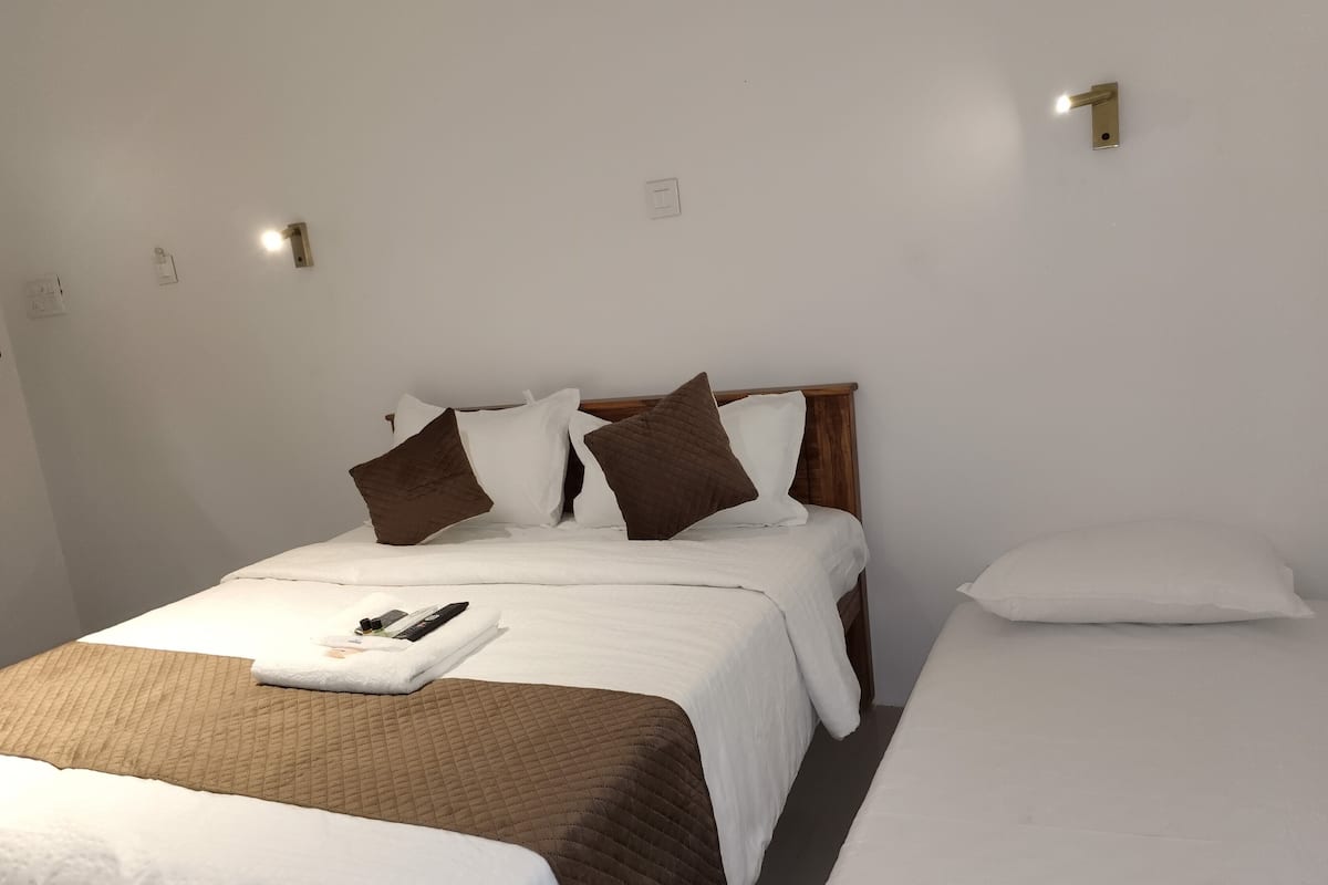 A double bed with a quilted brown bedspread and white pillows, a bedside table with a remote control, and a wall-mounted light fixture.