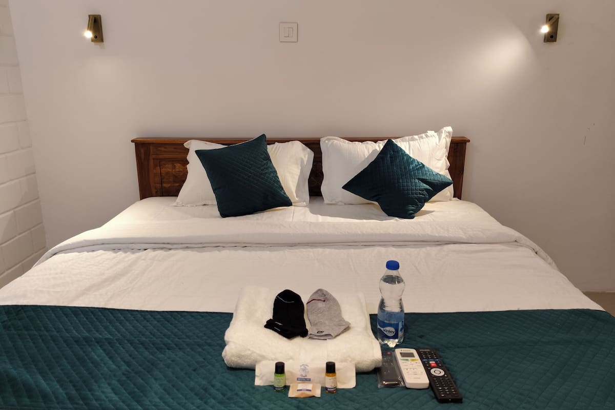 A neatly made bed with white and teal bedding, a teal quilted blanket, and a set of toiletries including a bottle of water, a remote control, and a cap.