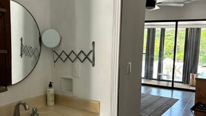 A bathroom with a large sink, a mirror, and a towel hanging on the wall. There is a window with curtains and a view of greenery outside. - 1BD Apartment with pool and beach access (Cancún)
