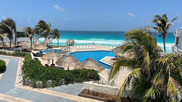 A resort pool area with a clear view of the ocean, surrounded by palm trees and thatched umbrellas. - 1BD Apartment with pool and beach access (Cancún)