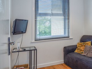 A living room with a flat-screen TV mounted on the wall, a glass coffee table, a sofa with patterned cushions, and a window with blinds.