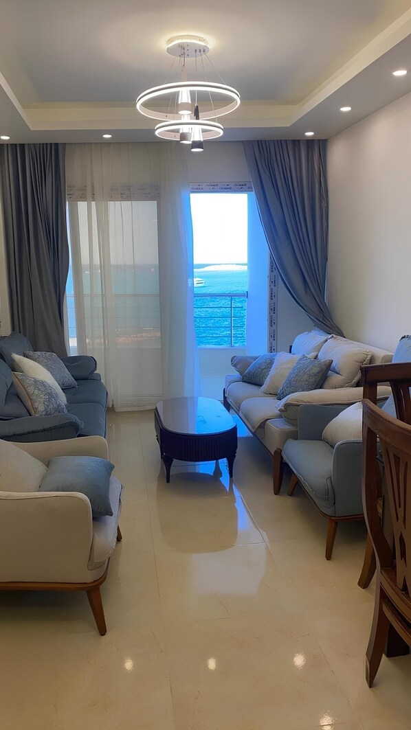 A spacious living room with two sofas, a coffee table, and a view of the ocean through a large window. - Al Emam Luxury Apartment (Marsa Matrouh)