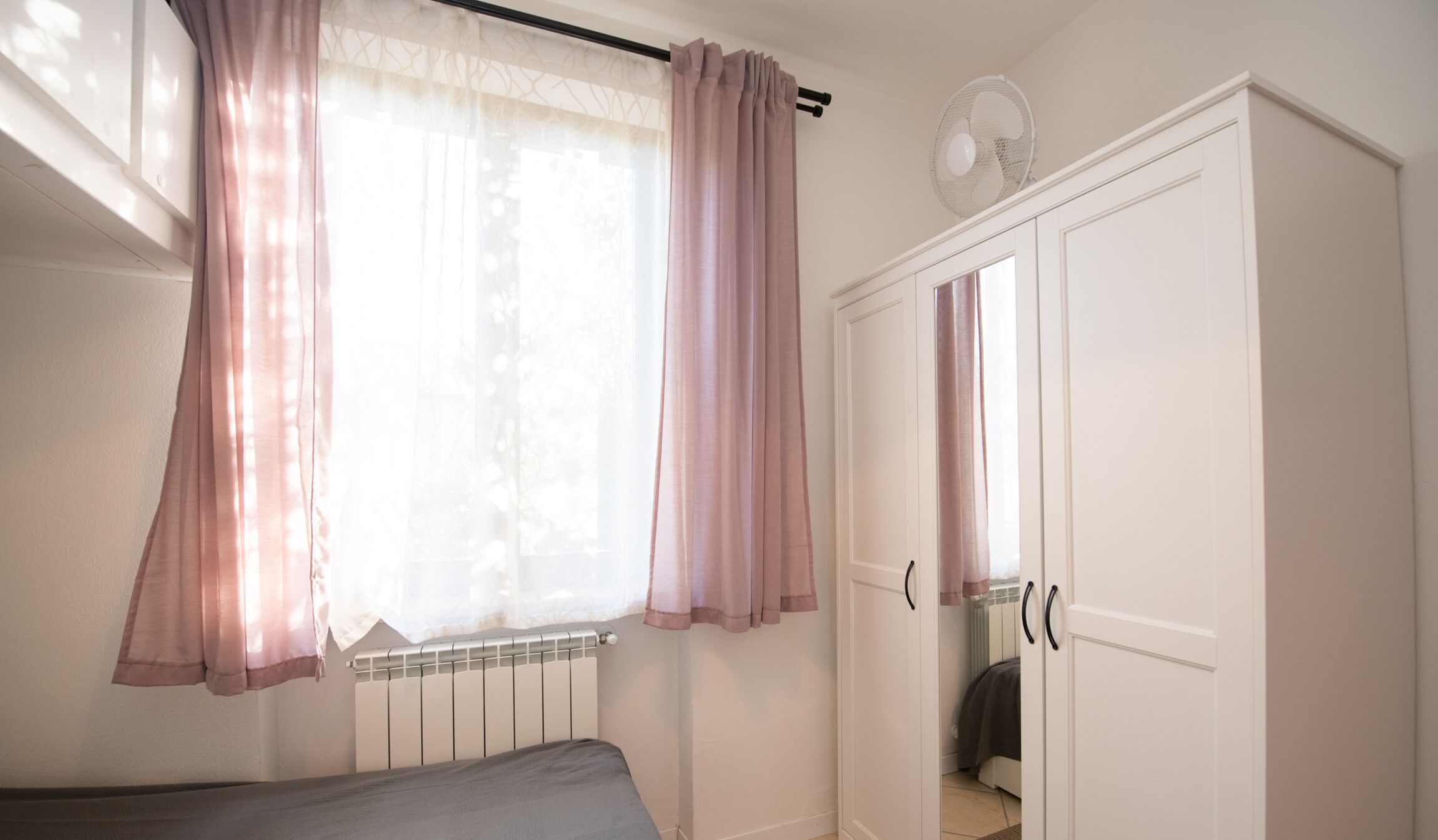 A bedroom with a bed, a radiator, a wardrobe, and a window with curtains.