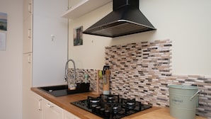 Une cuisine équipée d’une cuisinière à gaz, d’un évier et d’une hotte noire. Le mur derrière l’évier est recouvert de carreaux imitant des briques, et il y a un meuble blanc avec une poignée.