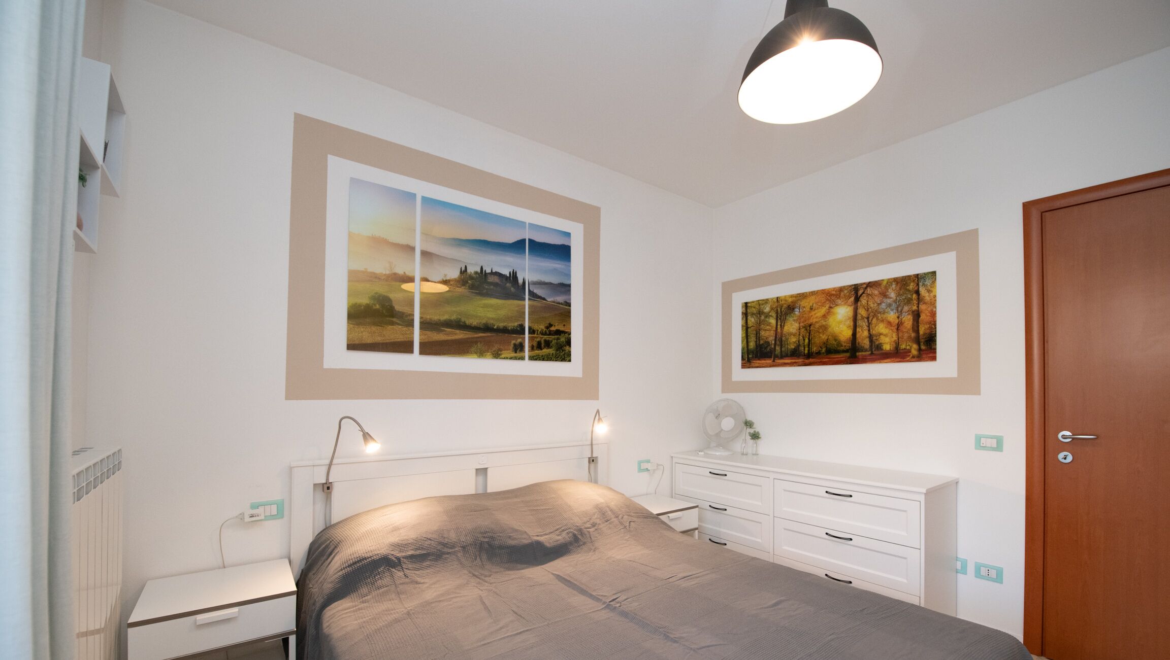 Une chambre à coucher comprenant un lit, des tables de chevet, une commode et deux tableaux représentant des paysages, encadrés.