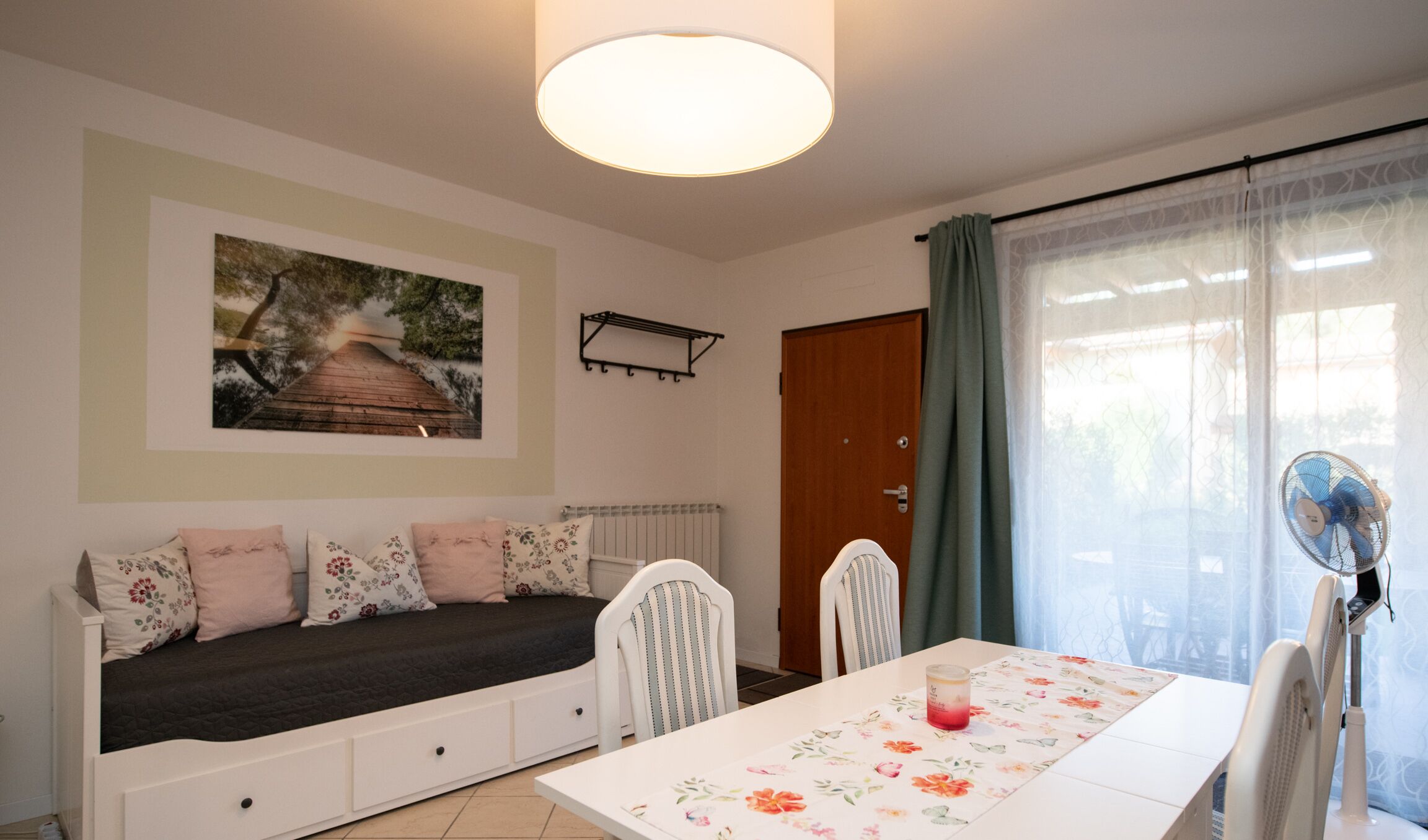 A cozy room with a bed, a dining table, chairs, a fan, a door, and a window with curtains.