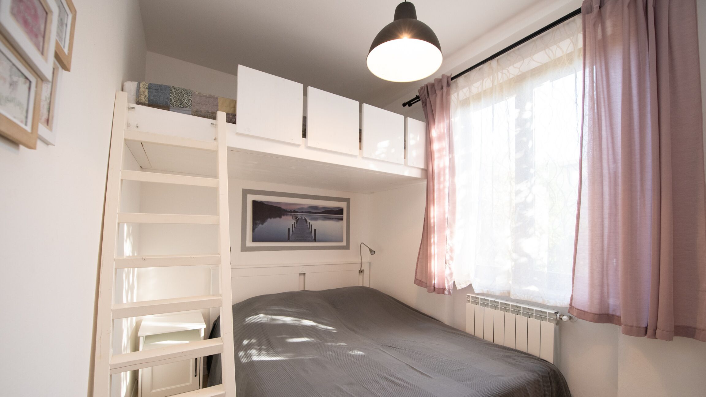 A bedroom with a bunk bed, a window with sheer curtains, a radiator, and a framed picture on the wall.
