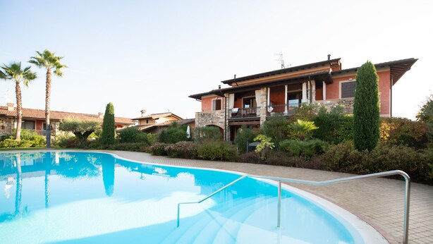 Une piscine à l’eau d’un bleu limpide, entourée d’une terrasse dallée. Au fond, on aperçoit un bâtiment à deux étages doté d’un balcon, d’une façade en pierre et d’un toit rouge.