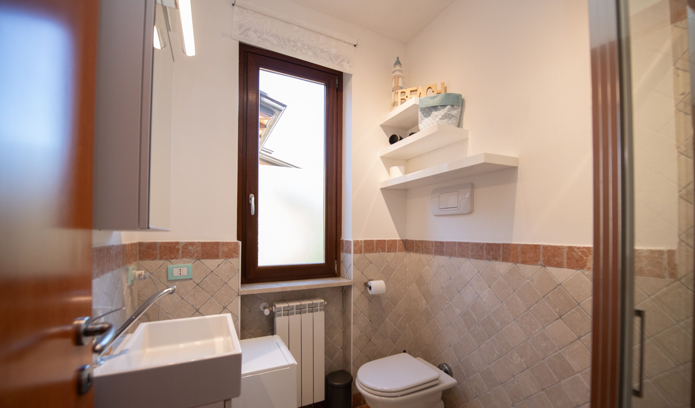 Ein Badezimmer mit Waschbecken, Toilette und einem Fenster. Über der Toilette befinden sich Ablagen mit Pflegeprodukten.
