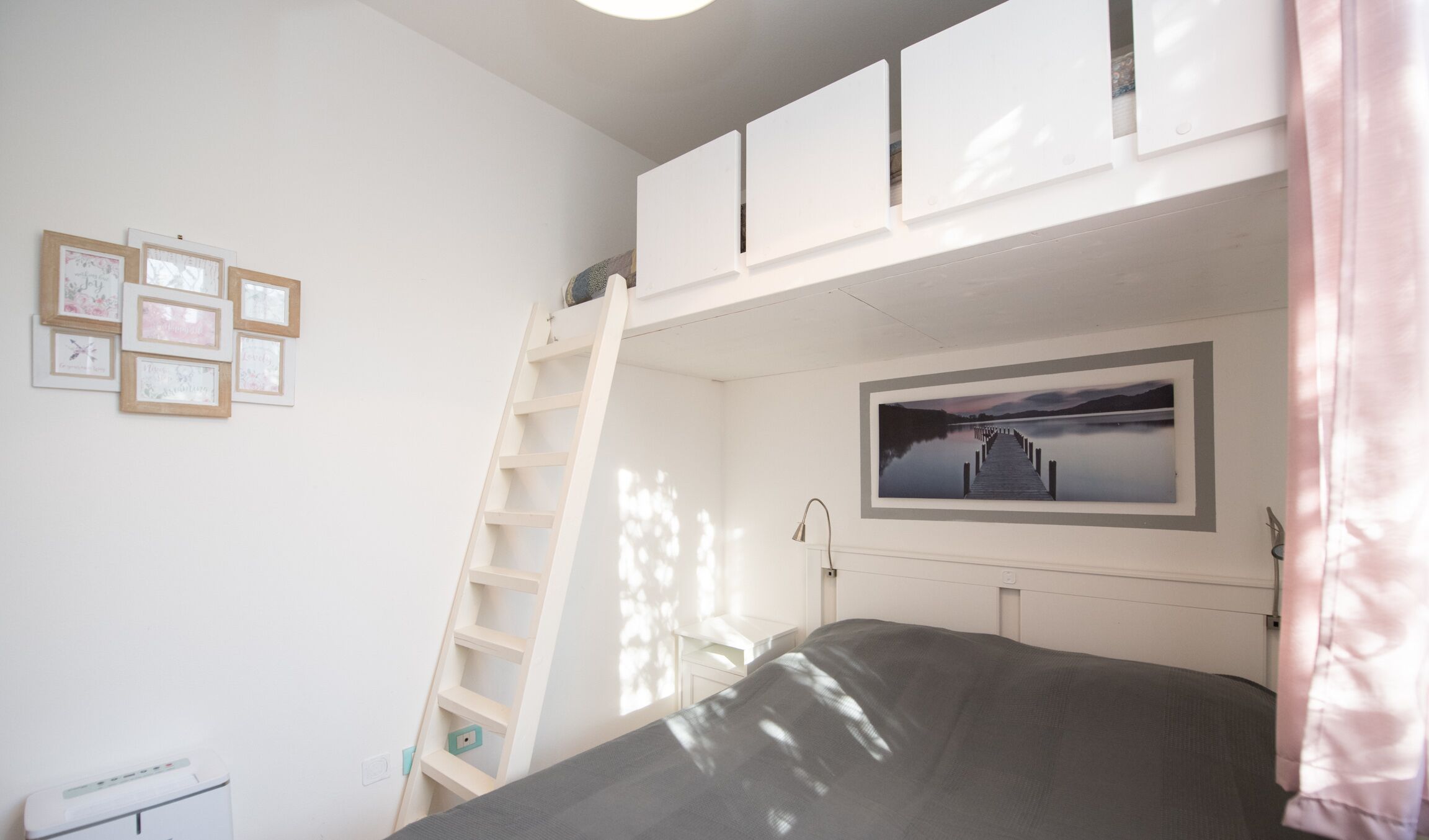 Une chambre avec un lit superposé, une échelle, une table de chevet, un tableau encadré et une fenêtre laissant entrer la lumière naturelle.