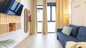 A modern living room with a flat-screen TV mounted on a wooden wall, a dark blue sofa with cushions, a small wooden shelf with a red object, and a large window with curtains.