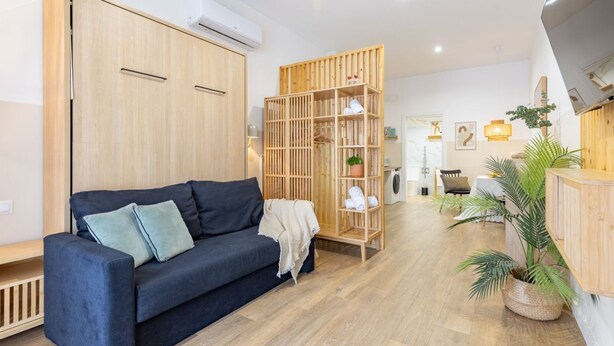A modern living room with a dark blue sofa, wooden wardrobe, and a wooden shelving unit with plants.
