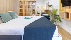 A modern hotel room with a bed, a wooden wardrobe, a desk with a chair, a TV mounted on the wall, and a potted plant.