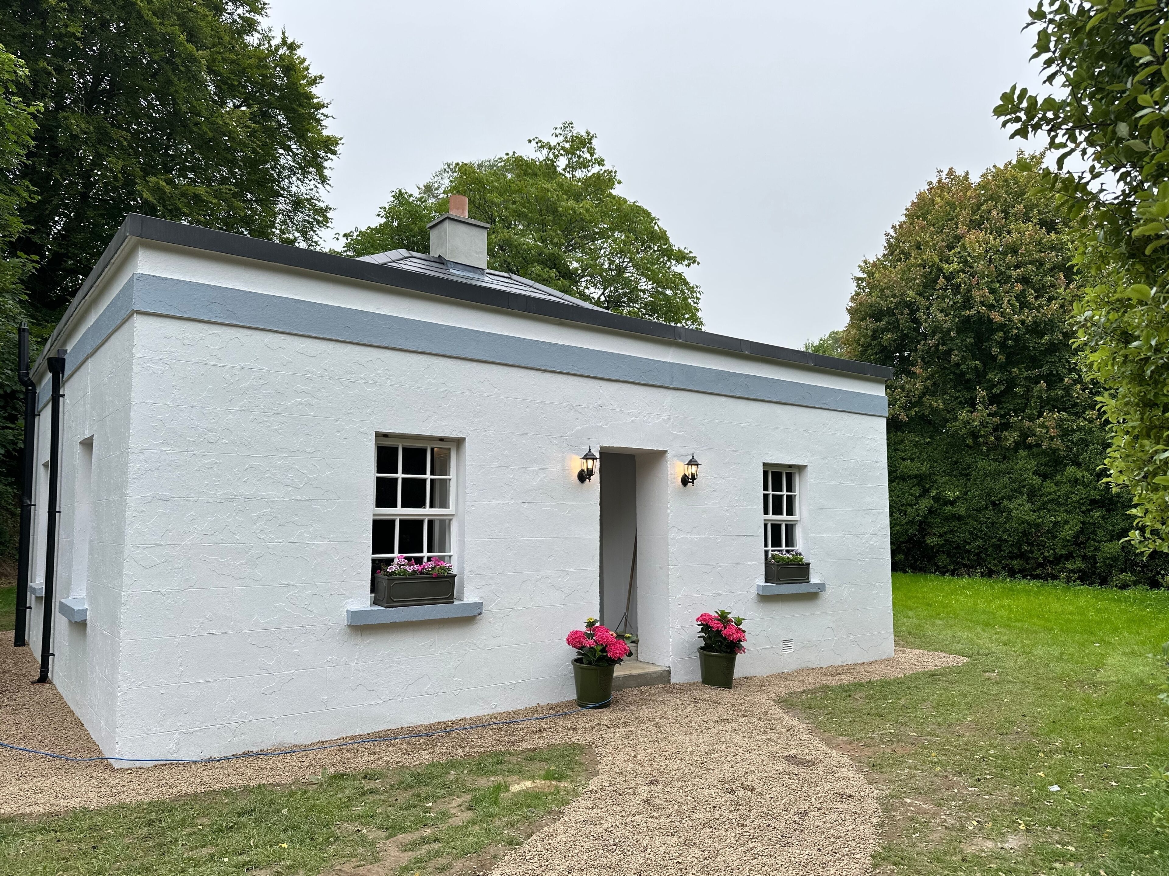 A small, white cottage with a grey roof, two windows with flower boxes, and a gravel path leading to the entrance.