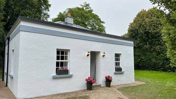 A small, white cottage with a grey roof, two windows with flower boxes, and a gravel path leading to the entrance.