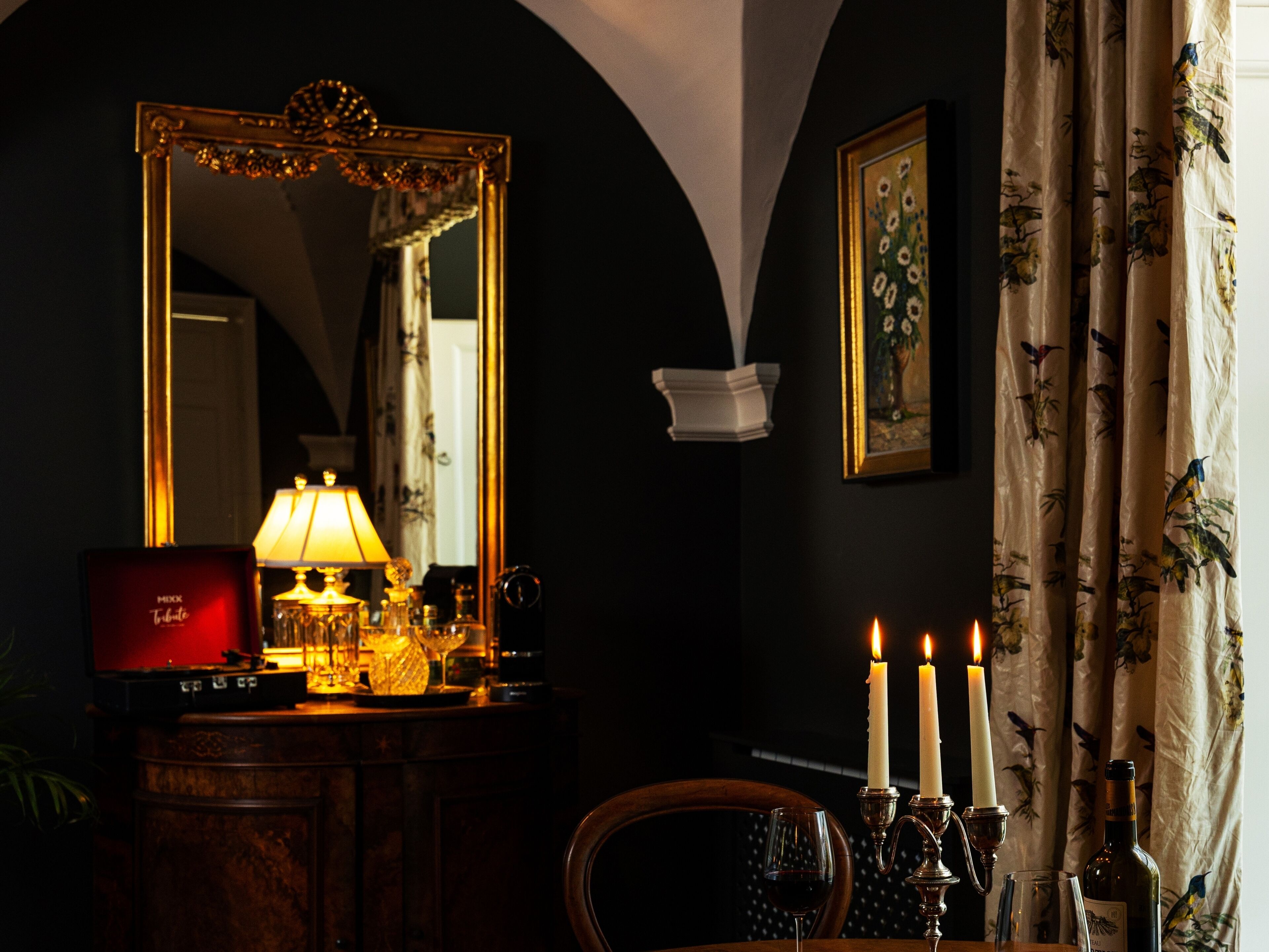 Une pièce faiblement éclairée, avec un grand miroir, une table sur laquelle se trouvent une lampe, des bougies et une bouteille.