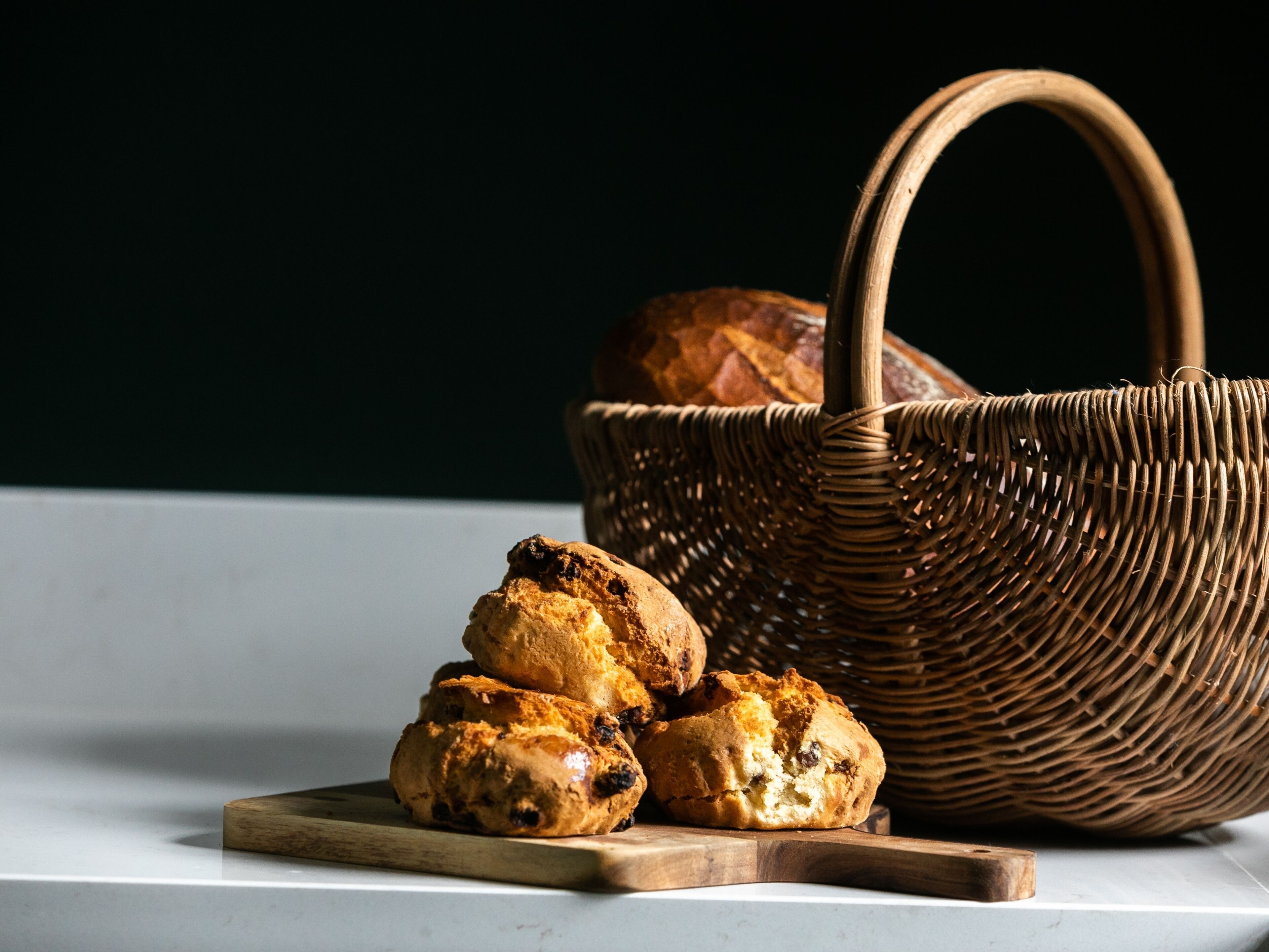 A wicker basket with a handle and a loaf of bread, alongside a stack of scones on a wooden board.