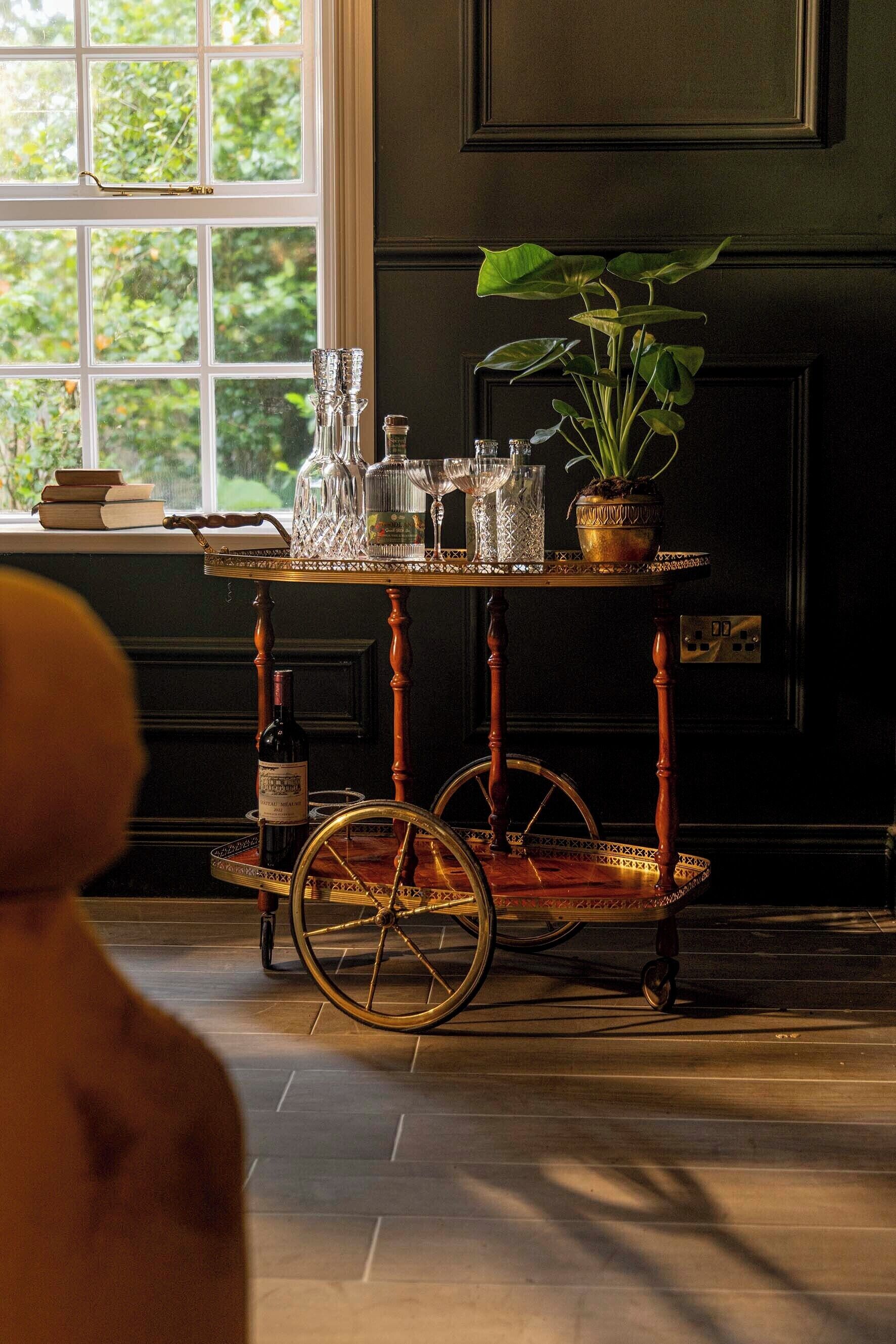 Une table de style vintage, agrémentée de verres et d’une plante en pot, placée près d’une fenêtre donnant sur un paysage verdoyant.