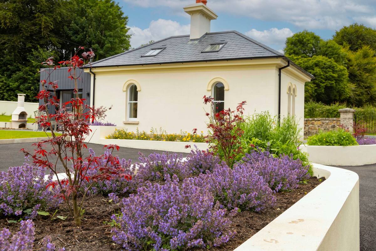 Een laag gebouw met een zadeldak en schoorsteen, omgeven door een aangelegde tuin met paarse bloemen en een witte steunmuur.
