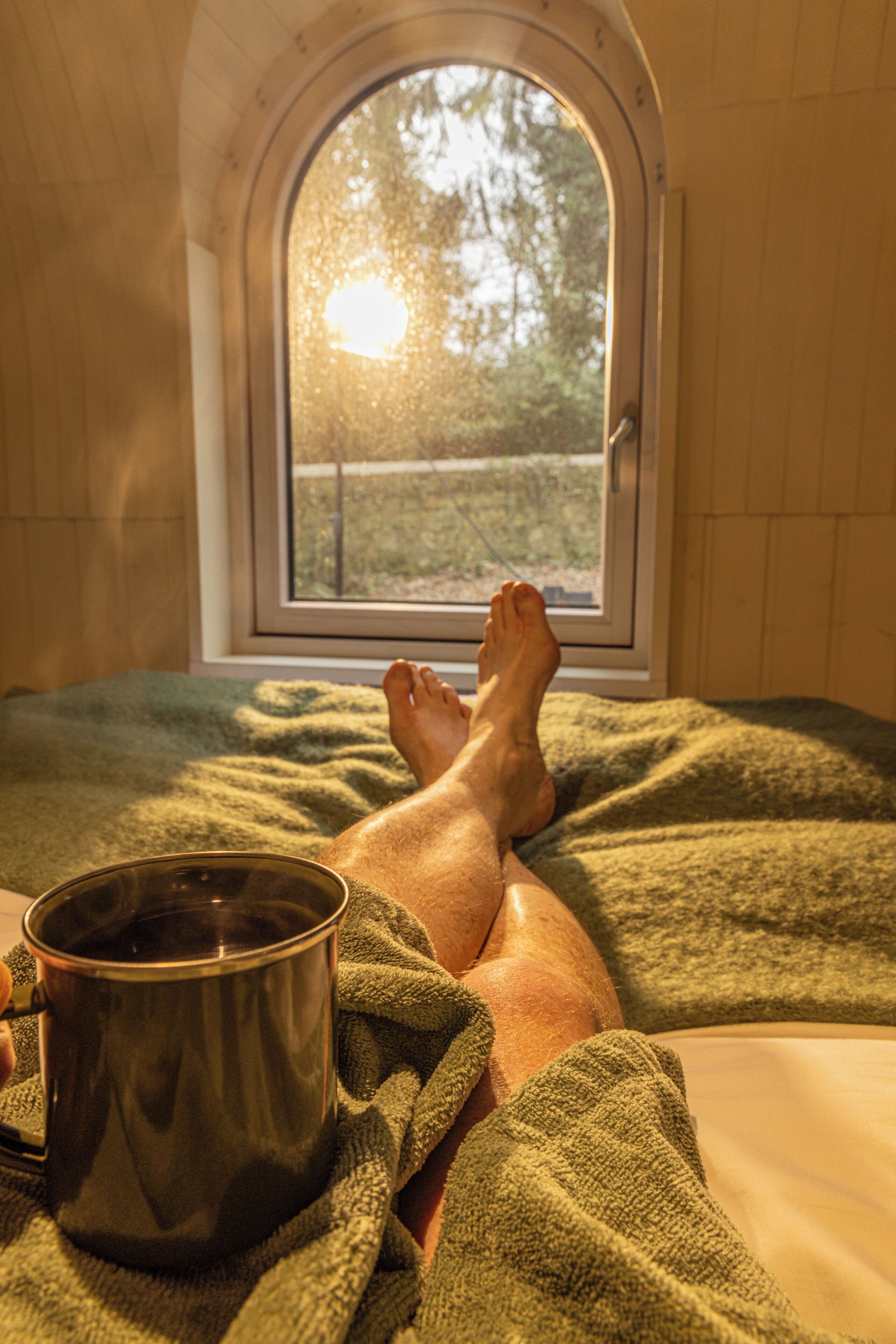 A person lying on a bed with a window behind them, a cup on the bed, and a green blanket.