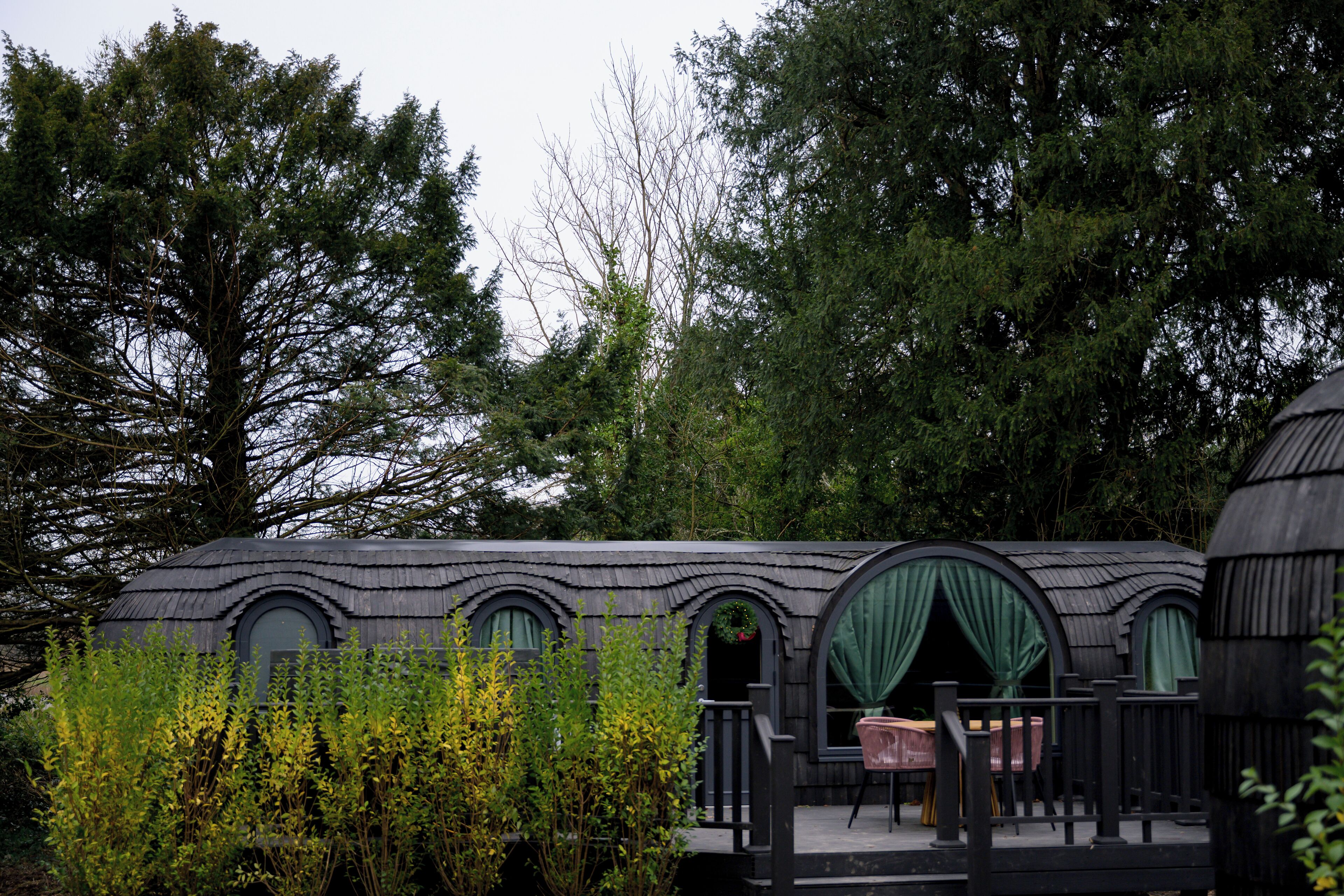 A rustic cabin with a curved roof, large windows with green curtains, and a wooden deck.
