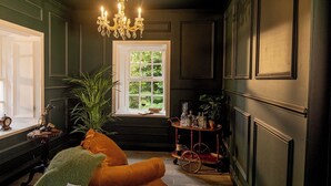 A room with dark walls, a chandelier, a window with a view of greenery, a wooden table with glassware, and a plush orange sofa. - The Forest at Mount Congreve Gardens (Waterford)