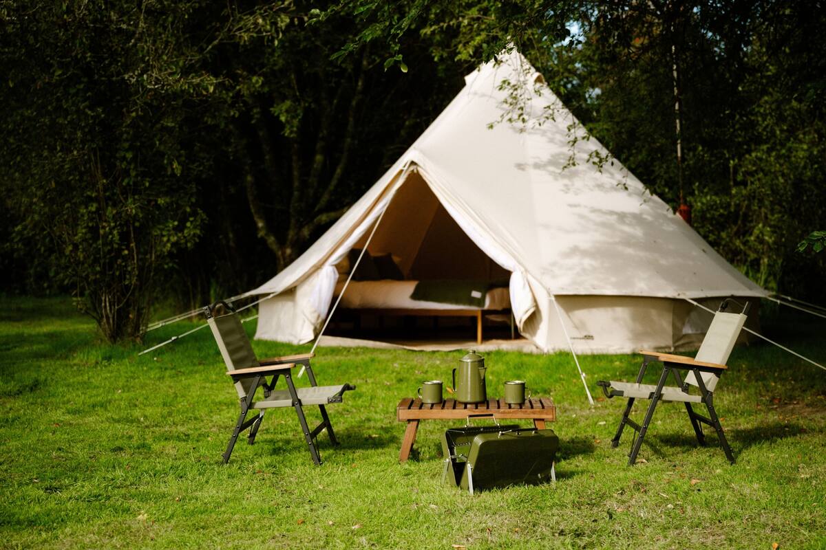 Een tent met een bed aan de binnenkant, omringd door stoelen en een tafel met bekers en een waterkoker.