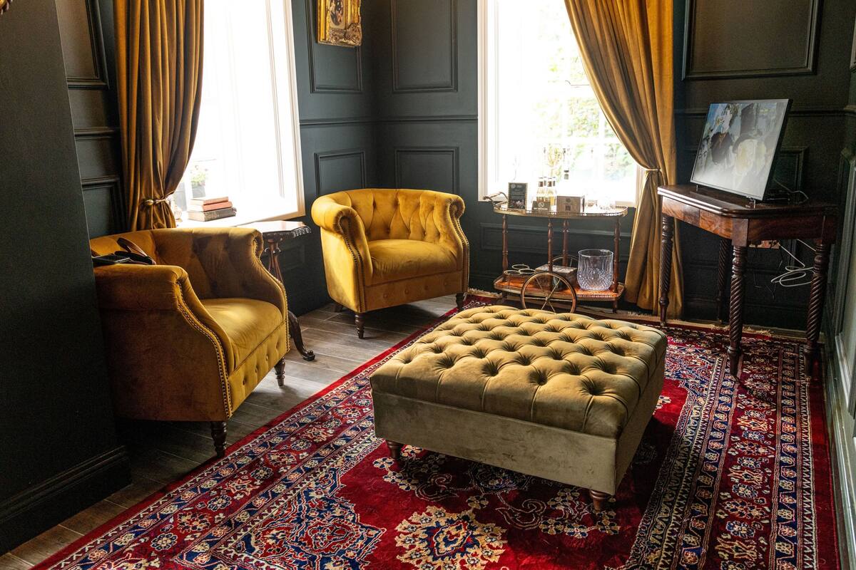 Een kamer met donkere muren, een gele fauteuil, een gestuufd poefje, een houten bijzettafeltje en een kamerbreed tapijt met een patroon.