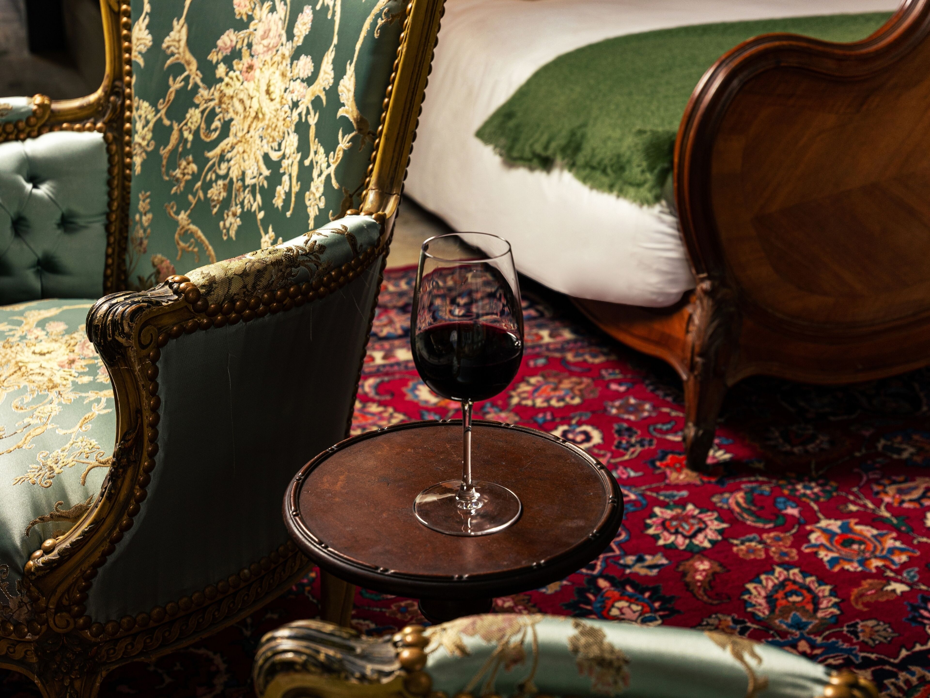 Ein Zimmer mit einem Bett, einem Sessel, einem Tisch mit einem Glas Rotwein und einem gemusterten Teppich.