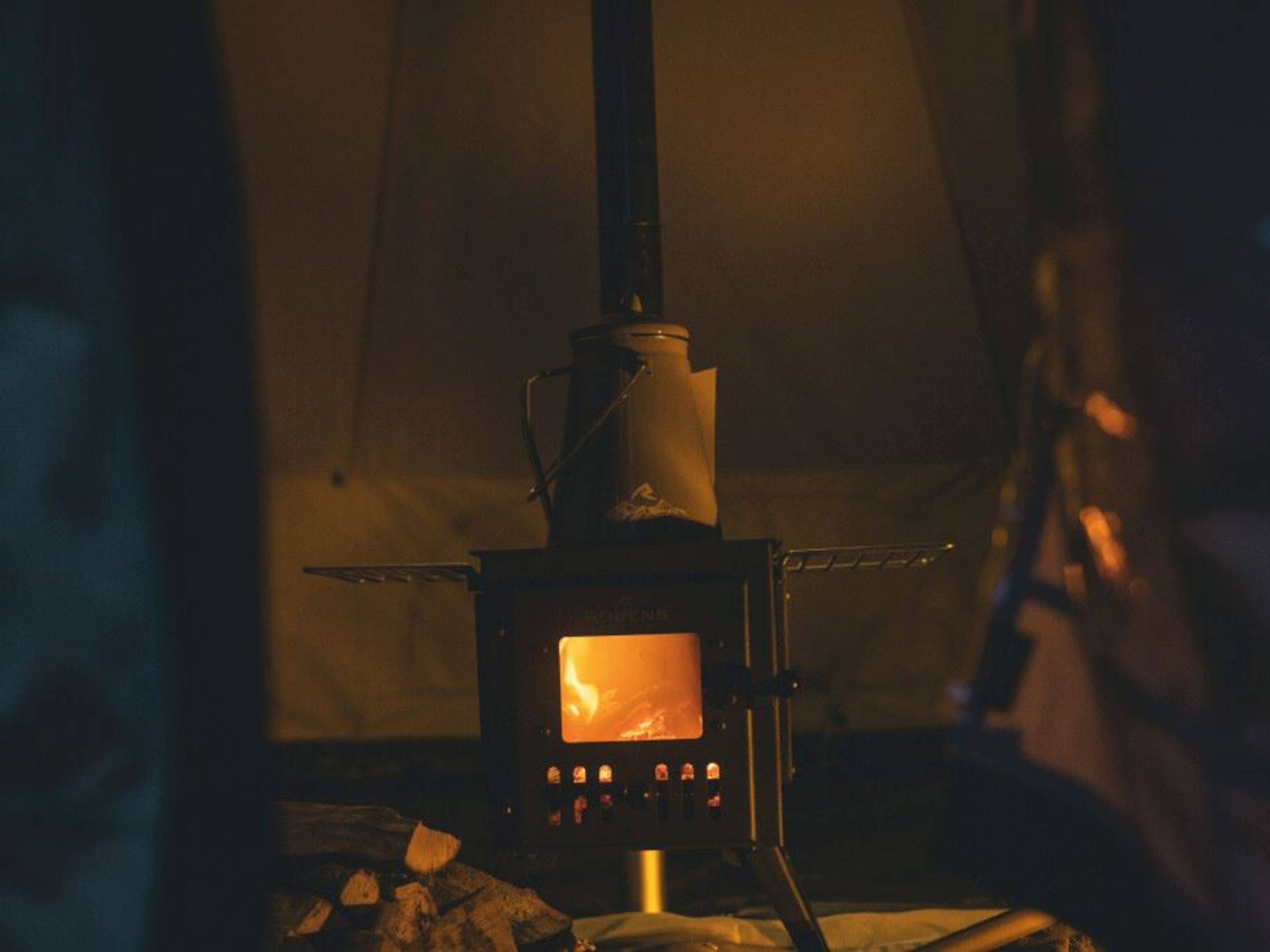 A wood-burning stove with a visible fire inside, placed in a room with a tent-like structure in the background.