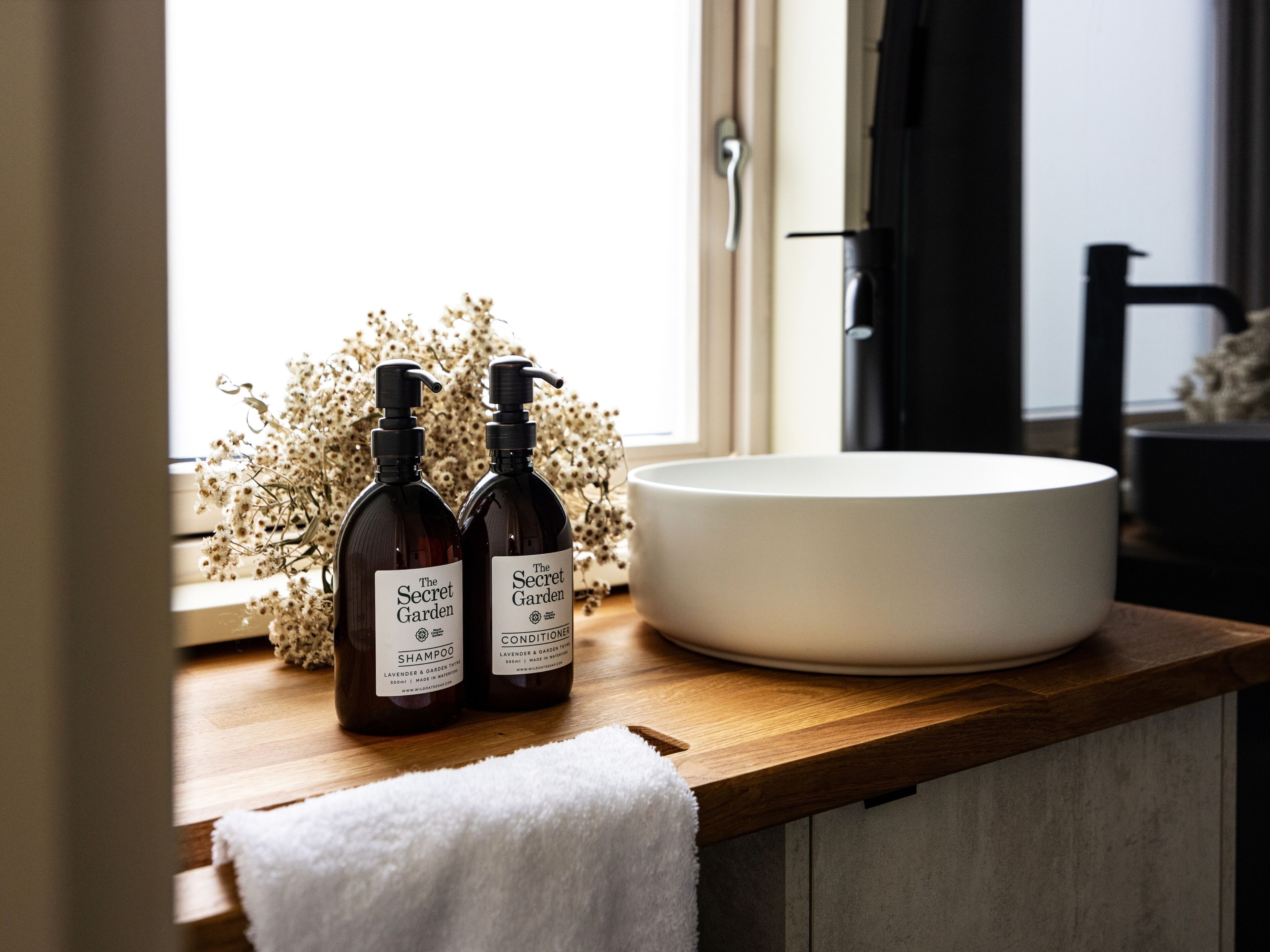 Ein Badezimmer-Waschtisch mit einer weißen Spüle, zwei Flaschen Shampoo und Conditioner von The Secret Garden und ein weißes Handtuch auf der Ablagefläche.