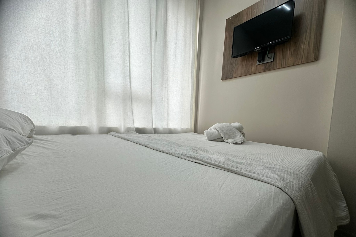 Une chambre d’hôtel avec un lit, un téléviseur fixé au mur et une fenêtre avec des rideaux.