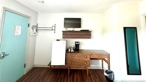 Una habitación funcional con escritorio de madera, televisor montado, refrigerador pequeño y perchero.