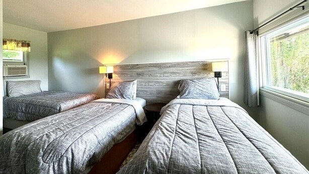 Habitación de hotel con dos camas individuales, cabecero de madera, ventana con cortinas y lámpara fijada a la pared.
