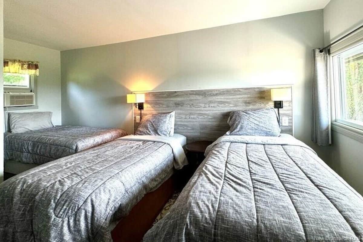 Une chambre d’hôtel avec deux lits simples, une tête de lit en bois, une fenêtre avec des rideaux et une lampe fixée au mur.