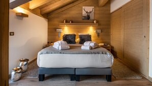 Ein gemütliches Schlafzimmer mit einem großen Bett, Holzbalken, einem Bild eines Reh und einem kleinen Nachttischchen mit einer Lampe.