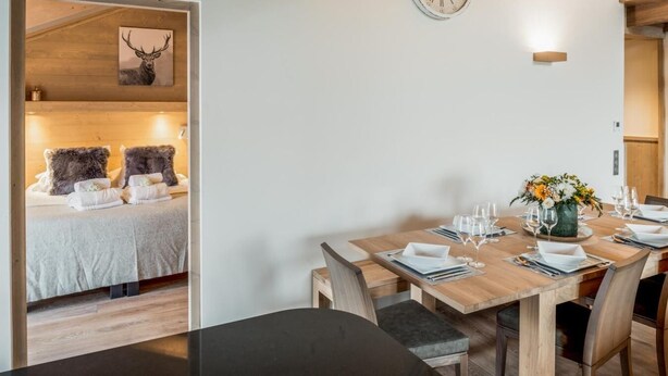 Ein modernes Zimmer mit einem Holztisch für vier Personen, einer Wanduhr und einem Bett mit einem Hirschgemälde.