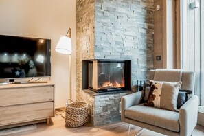 A cozy living room with a stone fireplace, a flat-screen TV, a comfortable armchair, and a wicker basket. - Wifi, 150m from ski slopes, 3rd floor, view mountain, sauna, balcony, fireplace or stove, parking (Courchevel)