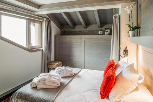 A bedroom with a bed, towels, a red pillow, a small stool, and a window.