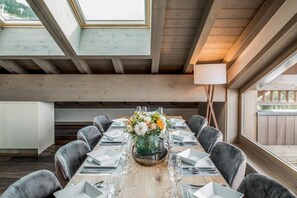A dining area with a long table set for six, surrounded by grey upholstered chairs. A vase with flowers is placed in the center. The room features exposed wooden beams and a large window on the right.