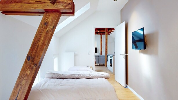 A bedroom with a sloped ceiling, a bed, a TV mounted on the wall, and a visible kitchen area in the background.