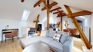 A modern loft apartment with exposed wooden beams, a large L-shaped gray sofa, a dining area, and a kitchen.