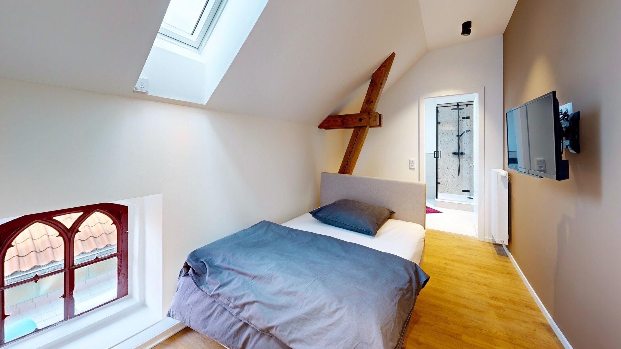 Ein Schlafzimmer mit Dachfenster, einem Bett mit grauer Bettdecke und Kissen, einem Holzkopfteil, einem an der Wand montierten Fernseher und einer Duschkabine aus Glas.