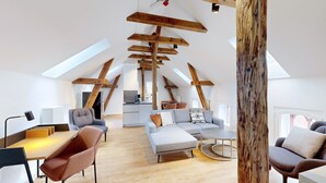 A modern loft apartment with exposed wooden beams, a dining area, a living room with a sofa and armchairs, and a kitchen in the background.